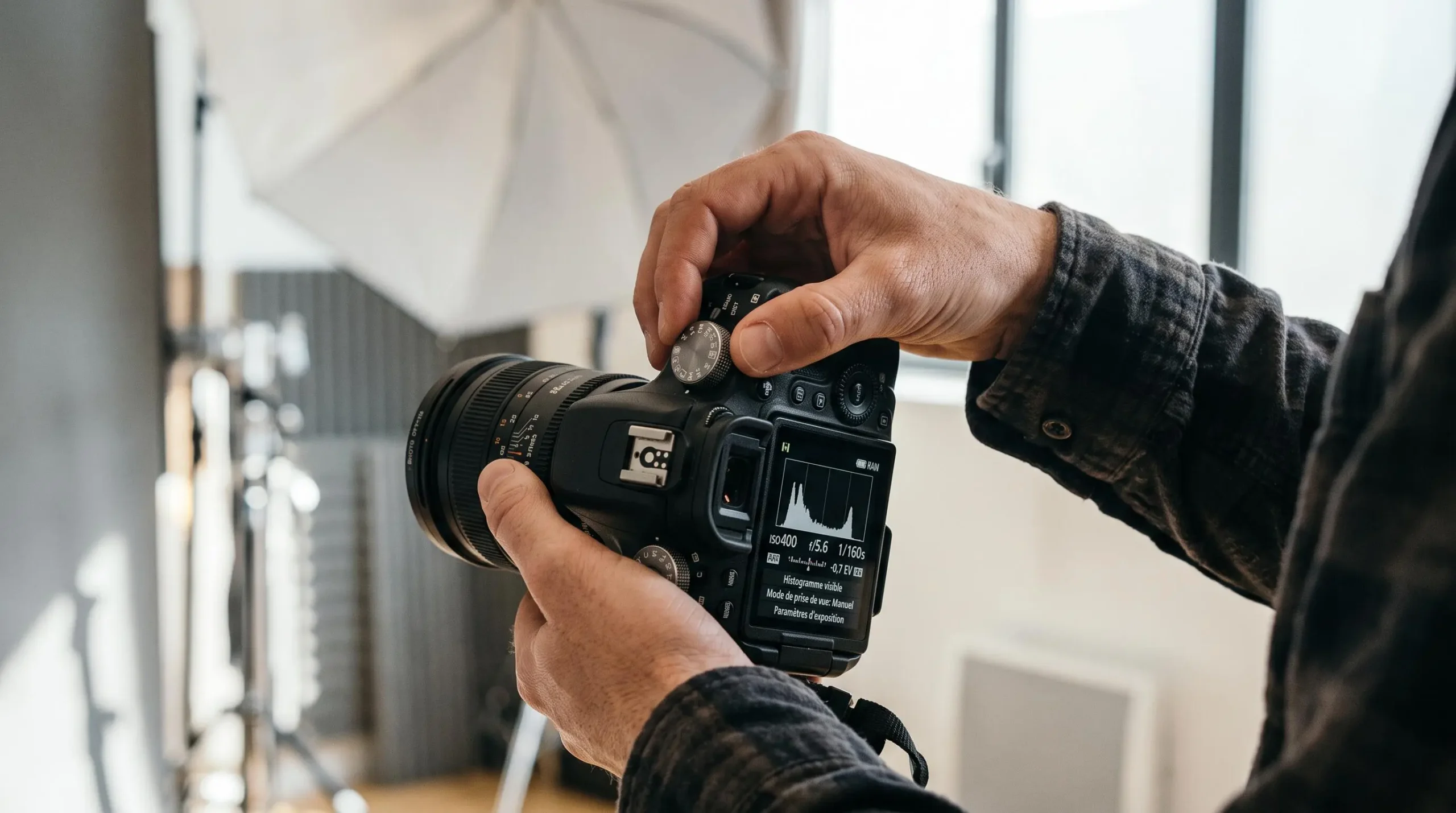 Gros plan sur les mains d'un photographe professionnel ajustant les réglages manuels d'un boîtier reflex plein format, écran LCD affichant histogramme et paramètres d'exposition